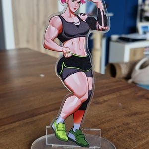 May include: A clear acrylic figure of a muscular woman flexing her arm. She wears a black sports bra, shorts with green trim, and green sneakers. The figure has pink hair and a tattoo on her arm. The figure is standing on a clear circular base.