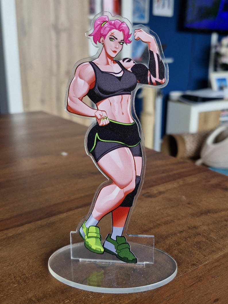 May include: A clear acrylic figure of a muscular woman flexing her arm. She wears a black sports bra, shorts with green trim, and green sneakers. The figure has pink hair and a tattoo on her arm. The figure is standing on a clear circular base.