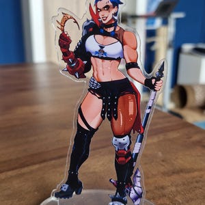 May include: A clear acrylic figure of a female character with blue hair, wearing a white and black top, black shorts, and knee-high boots. She is holding a weapon and has a determined expression. The figure is mounted on a clear circular base.