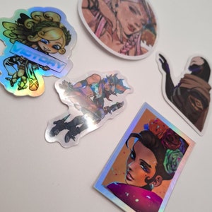 May include: Five holographic stickers featuring various illustrations. One sticker features a woman with pink hair and a floral crown. Another sticker features a woman with brown hair and a floral crown. A third sticker features a woman with green hair and wings. A fourth sticker features a woman with blue hair and a sword. The fifth sticker features a person with a black cloak and a hood.