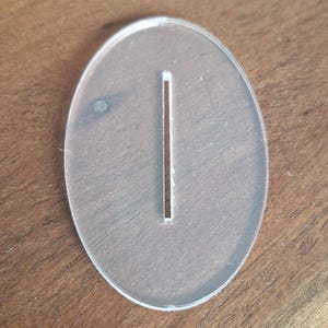 May include: A clear, oval-shaped acrylic base featuring a single, centered, vertical rectangular slot. The piece is smooth, transparent, and flat, designed with a clean edge profile and a uniform thickness throughout its entire structure.
