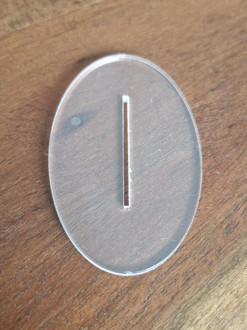 May include: A clear, oval-shaped acrylic base featuring a single, centered, vertical rectangular slot. The piece is smooth, transparent, and flat, designed with a clean edge profile and a uniform thickness throughout its entire structure.