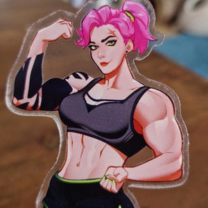 May include: A clear acrylic sticker featuring a muscular woman with pink hair in a ponytail, wearing a black sports bra and shorts with green trim. The woman is flexing her arm, showcasing her strength.