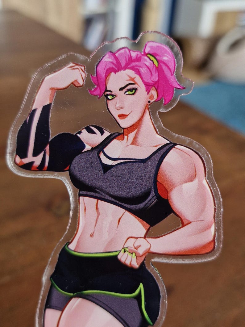 May include: A clear acrylic sticker featuring a muscular woman with pink hair in a ponytail, wearing a black sports bra and shorts with green trim. The woman is flexing her arm, showcasing her strength.