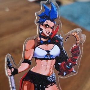 May include: A clear acrylic charm featuring a character with blue spiky hair, wearing a white crop top with a skull emblem, black shorts, and a spiked gauntlet on one arm. The character holds a long staff in one hand and a curved blade in the other.