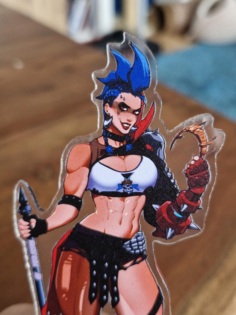 May include: A clear acrylic charm featuring a character with blue spiky hair, wearing a white crop top with a skull emblem, black shorts, and a spiked gauntlet on one arm. The character holds a long staff in one hand and a curved blade in the other.