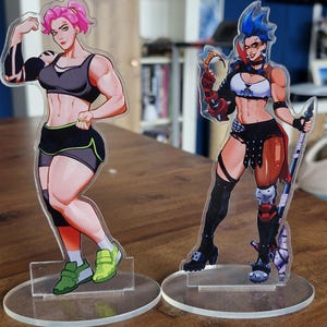 May include: Two acrylic character standees featuring illustrated figures. The left figure has pink hair, a black sports bra, and black shorts. The right figure has blue hair, a white crop top, black shorts, and holds a spear-like weapon.