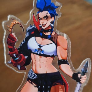 May include: A clear acrylic standee featuring a character with bright blue hair, wearing a white crop top with a skull emblem, black shorts, and red spiked arm armor. The character holds a curved golden blade in one hand and a spear in the other.