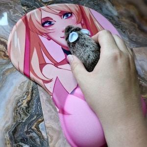May include: A pink and white anime-style mousepad with a female character with long blonde hair. A small gray and brown plush mouse is on the mousepad.
