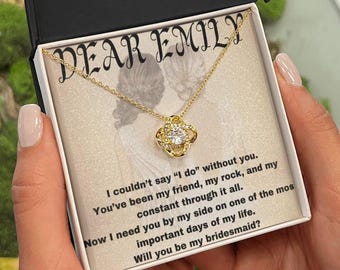 May include: A gold-colored necklace with a four-leaf clover pendant, presented in a gift box. The box lid is open, revealing the necklace and a card with the text "DEAR EMILY" and a bridesmaid proposal.