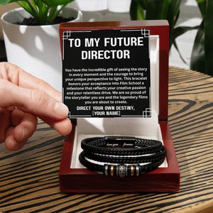 May include: A black leather bracelet with silver and gold accents is displayed inside a red wooden box. The box lid has text that reads "TO MY FUTURE DIRECTOR". The bracelet is a gift for someone in film school.