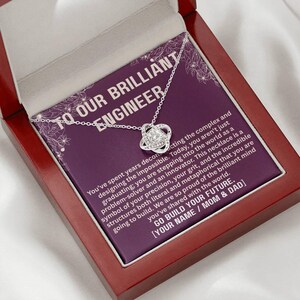 May include: A silver-colored necklace with a knot pendant, presented in a maroon gift box. The box contains a purple card with white text that reads "TO OUR BRILLIANT ENGINEER" and a message. A thoughtful gift.