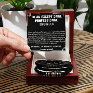 May include: A black braided leather bracelet with silver and gold accents, presented in a dark wooden box. The box contains a message for a professional engineer. The bracelet has the words "love you forever" engraved on it.