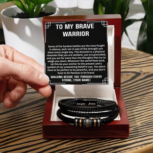 May include: A black braided bracelet with silver and gold accents, presented in a dark wooden box. The box contains a card with the text "TO MY BRAVE WARRIOR" and a message of support. The bracelet is a thoughtful gift.