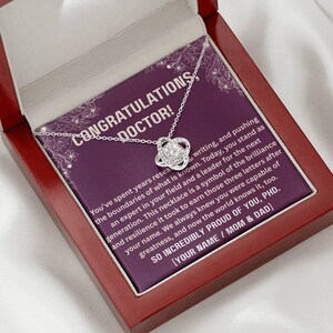 May include: A silver necklace with a knot pendant, presented in a red wooden box. The box contains a purple card with the text "CONGRATULATIONS, DOCTOR!" and a message. The necklace is a thoughtful gift for a new doctor.