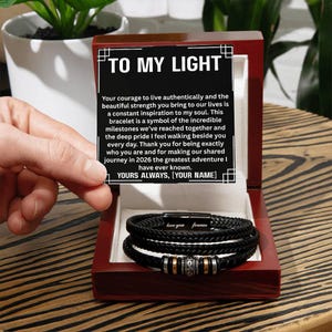 May include: A black leather bracelet with silver and gold accents, presented in a wooden box. The box lid displays the text "TO MY LIGHT" and a heartfelt message. The bracelet is inscribed with "love you forever".