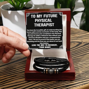 May include: A black leather bracelet with silver and gold accents, presented in a wooden box. The box contains a message for a future physical therapist, with the text "TO MY FUTURE PHYSICAL THERAPIST". The bracelet is inscribed with "love you forever".
