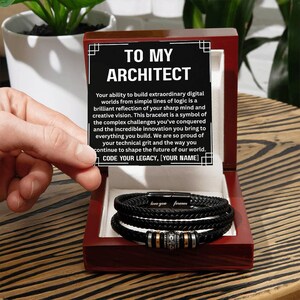 May include: A black braided bracelet with silver and gold accents, presented in a dark red wooden box. The box contains a card with the text "TO MY ARCHITECT" and a message. The bracelet is a thoughtful gift.