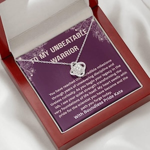 May include: A silver necklace with a knot pendant, presented in a red wooden box. The box contains a purple card with white text that reads "TO MY UNBEATABLE WARRIOR" and a message about reaching a milestone.