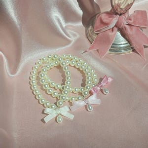 May include: Three pearl bracelets with delicate bows in white, pink, and light pink. The bracelets are on a pink satin background.