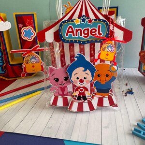 May include: A clear plastic gift bag with a circus-themed design. The bag features a red and white striped tent, the name "Angel", and cartoon characters. The bag is decorated with red, white, blue, and yellow accents.