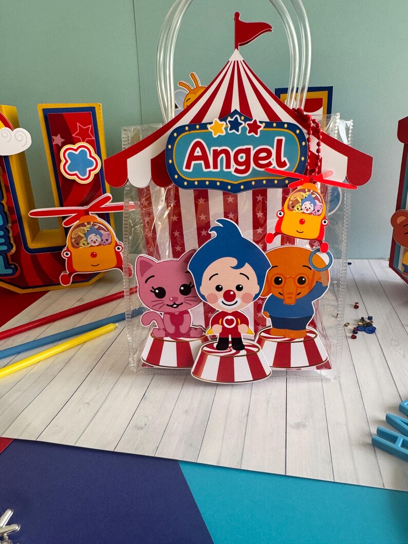 May include: A clear plastic gift bag with a circus-themed design. The bag features a red and white striped tent, the name "Angel", and cartoon characters. The bag is decorated with red, white, blue, and yellow accents.