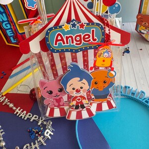 May include: A clear plastic gift bag with a red and white circus tent design, the name "Angel" and cartoon characters. The bag has a clear handle and is filled with treats. Other party decorations are in the background.