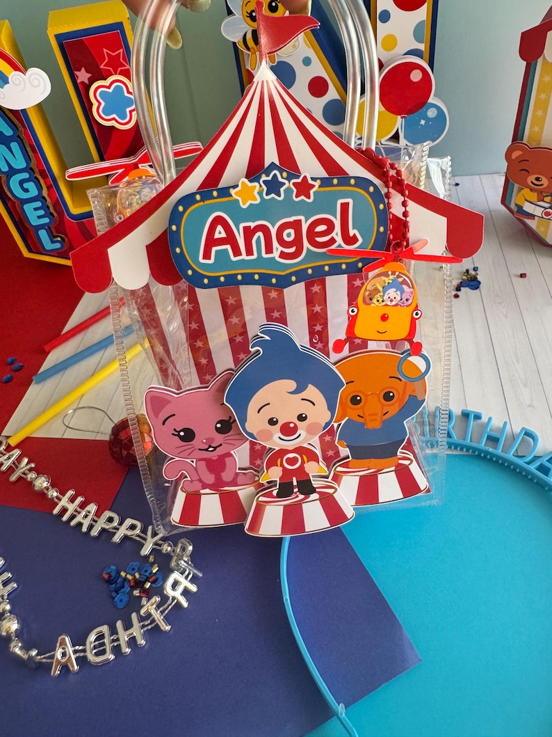 May include: A clear plastic gift bag with a red and white circus tent design, the name "Angel" and cartoon characters. The bag has a clear handle and is filled with treats. Other party decorations are in the background.
