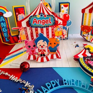May include: A colorful birthday party scene with a circus theme. A clear plastic bag with a red and white striped circus tent design and the name "Angel" is in the center. Other decorations include a rainbow, a "Happy Birthday" headband, and a "Happy" necklace.