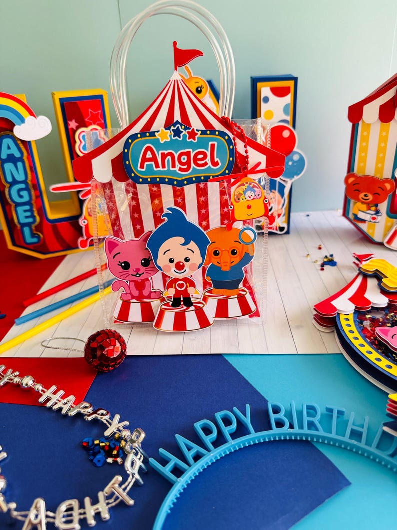 May include: A colorful birthday party scene with a circus theme. A clear plastic bag with a red and white striped circus tent design and the name "Angel" is in the center. Other decorations include a rainbow, a "Happy Birthday" headband, and a "Happy" necklace.