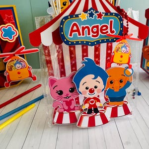 May include: A clear plastic gift bag with a circus tent design, featuring the name "Angel" in red letters. The bag includes cartoon characters and a red, white, and blue color scheme. Additional decorative elements include a helicopter and colorful pencils.