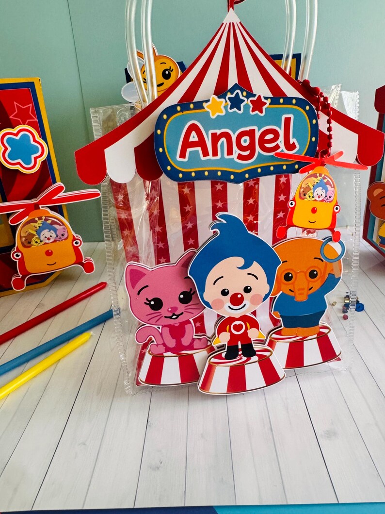 May include: A clear plastic gift bag with a circus tent design, featuring the name "Angel" in red letters. The bag includes cartoon characters and a red, white, and blue color scheme. Additional decorative elements include a helicopter and colorful pencils.