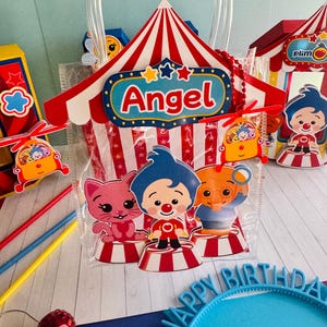 May include: A clear plastic party favor bag with a red and white circus tent design, the name "Angel" and cartoon characters. The bag has a clear handle and is filled with party favors. Other circus-themed decorations are visible.
