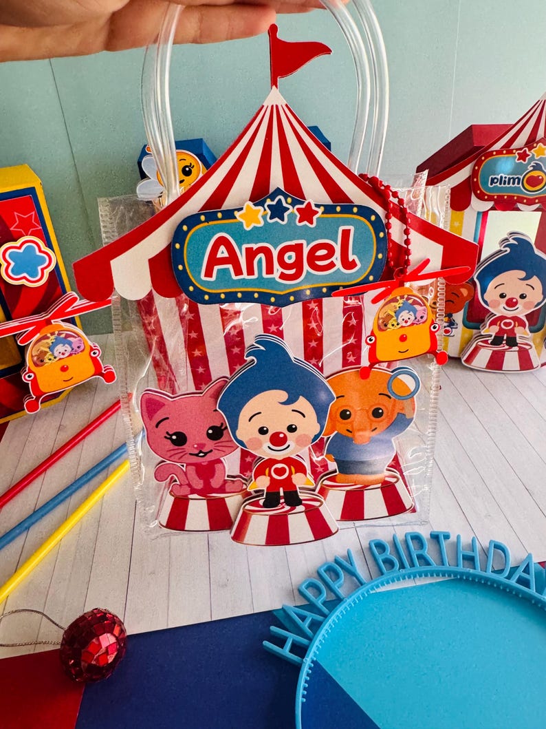 May include: A clear plastic party favor bag with a red and white circus tent design, the name "Angel" and cartoon characters. The bag has a clear handle and is filled with party favors. Other circus-themed decorations are visible.