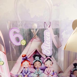 Puede incluir: Bolsa de regalo de plástico transparente con un diseño temático de spa. La bolsa presenta un tipi con tres chicas de dibujos animados con batas de spa. Se adjunta un número 6 morado y el nombre "Alexa". También se incluye un dije en forma de peine.