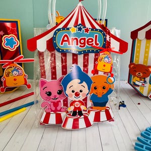 May include: A clear plastic gift bag with a circus theme. The bag features a red and white striped tent design with the name "Angel" on it. The bag is decorated with cartoon characters and a red flag.