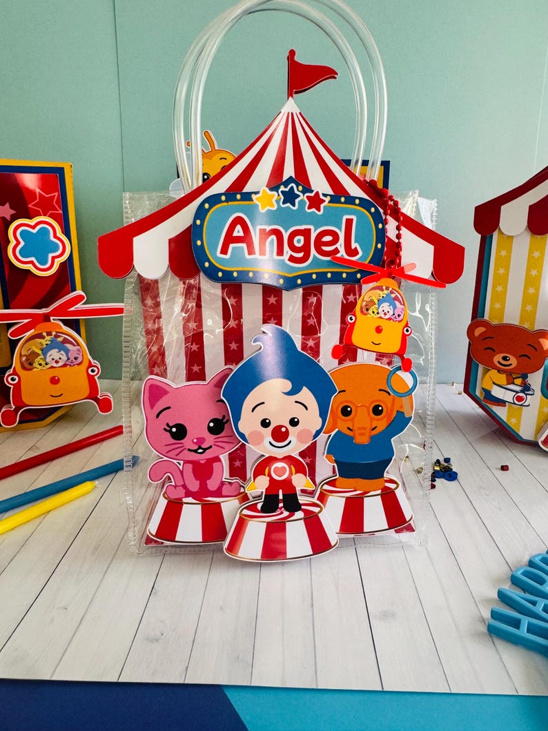 May include: A clear plastic gift bag with a circus theme. The bag features a red and white striped tent design with the name "Angel" on it. The bag is decorated with cartoon characters and a red flag.