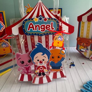May include: A clear plastic gift bag with a circus tent design. The tent is red and white striped with the name "Angel" on a blue sign. The bag features cartoon characters of a cat, clown, and elephant on red and white striped platforms.