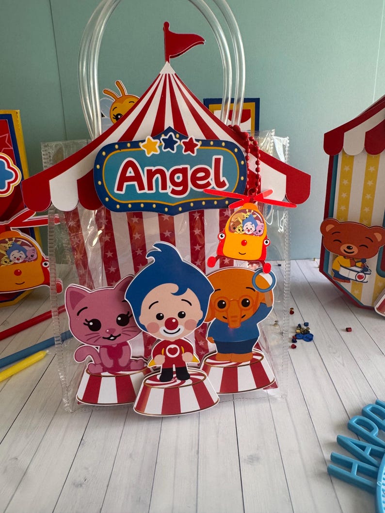 May include: A clear plastic gift bag with a circus tent design. The tent is red and white striped with the name "Angel" on a blue sign. The bag features cartoon characters of a cat, clown, and elephant on red and white striped platforms.