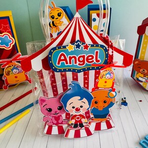 May include: Clear plastic gift bag with a circus theme. The bag features a red and white striped circus tent design with the name "Angel" on it. Inside are cartoon characters of a cat, clown, and elephant. A yellow bee is also visible.