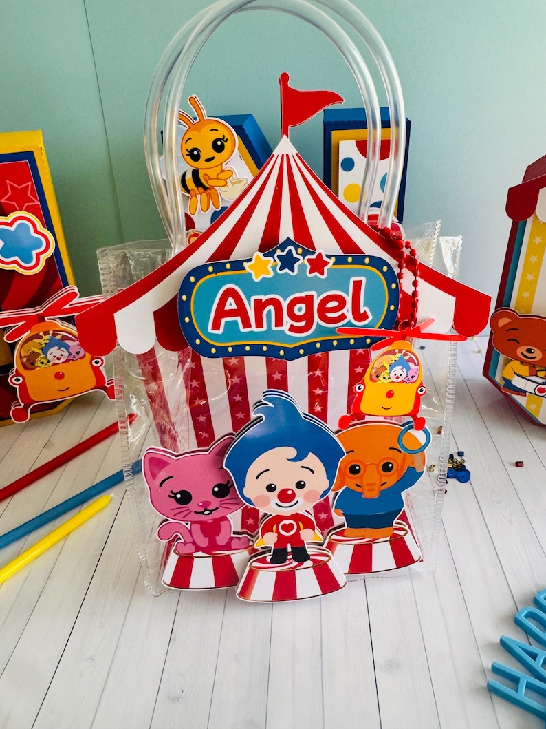 May include: Clear plastic gift bag with a circus theme. The bag features a red and white striped circus tent design with the name "Angel" on it. Inside are cartoon characters of a cat, clown, and elephant. A yellow bee is also visible.