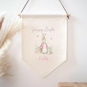 May include: A white canvas banner with the text "Happy Easter" and a watercolour illustration of a pink bunny rabbit with flowers. The banner also has the name "Lola" printed on it.