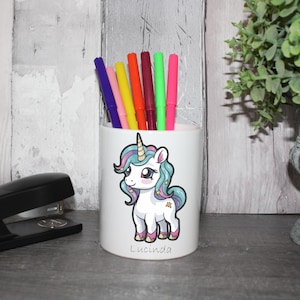 May include: White ceramic pencil holder featuring a cartoon unicorn design. The unicorn has a blue and pink mane and tail, and is holding a gold star. The pencil holder is personalised with the name "Lucinda".