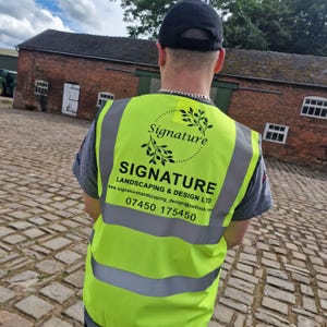 Puede incluir: Una persona lleva un chaleco de seguridad amarillo con el texto "SIGNATURE LANDSCAPING & DESIGN LTD" y el n&uacute;mero de tel&eacute;fono "07450 175450". El chaleco tambi&eacute;n tiene un logotipo con la palabra "Signature" en un c&iacute;rculo con un dise&ntilde;o de hojas.