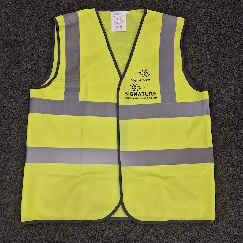Puede incluir: Un chaleco de seguridad amarillo brillante con rayas reflectantes plateadas. El chaleco tiene un logotipo negro con el texto "Signature Landscaping & Design LTD".