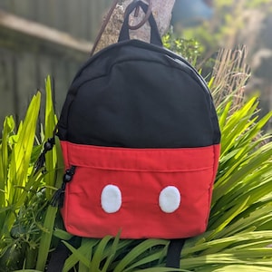May include: A black and red backpack with white circular accents, reminiscent of a cartoon character's face. The backpack has a top handle and adjustable straps. The bag is hanging outside in front of green foliage.