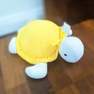 May include: A plush turtle toy featuring a bright yellow shell and white head, limbs, and tail. A yellow satin ribbon is tied around the neck of the turtle. A small white rectangular product tag is attached to the ribbon near the turtle's head.