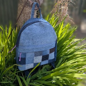 May include: A denim backpack in shades of blue, featuring a patchwork design on the front. It has a top handle, zip closure, and adjustable straps. The bag is suspended from a metal ring, set against a natural backdrop.