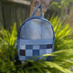 May include: A denim backpack with a patchwork design on the lower front. The backpack is a mix of light and dark blue denim, with a small handle at the top. The bag is hanging from a tree branch, with green foliage in the background.