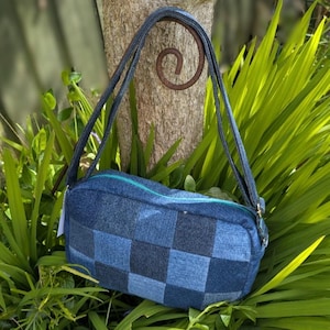 May include: A blue denim patchwork handbag with a rectangular shape and a teal zip. The bag has long, thin straps. The denim is a mix of dark and light blue squares. The bag is set against a backdrop of greenery and a tree trunk.